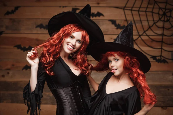 Концепция Хэллоуина - красивая кавказская мать и ее дочь с длинными рыжими волосами в костюмах ведьм празднуют Хэллоуин позируя с над летучими мышами и паутиной на фоне студии Wooden . — стоковое фото