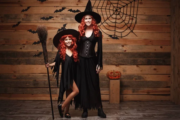 Halloween Concept - mooie Kaukasische moeder en haar dochter met lange rode haren in heks kostuums vieren Halloween poseren met over vleermuizen en spinnenweb op houten studio achtergrond. — Stockfoto