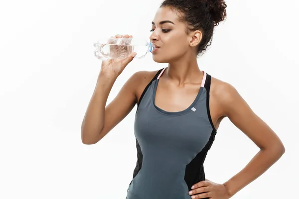 Conceito saudável e Fitness - bela menina afro-americana em roupas esportivas beber água por garrafa de plástico após o treino. Isolado no fundo do estúdio branco — Fotografia de Stock