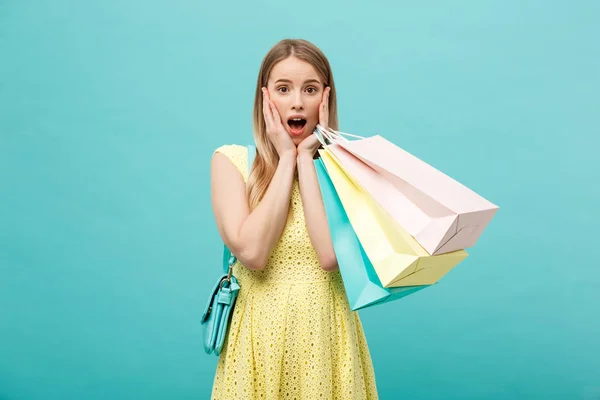 Lifestylekonzept: Porträt schockiert junge brünette Frau in gelbem Sommerkleid posiert mit Einkaufstaschen vor pastellblauem Hintergrund — Stockfoto