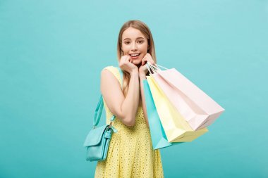 Yaşam Tarzı Konsepti: Sarı yaz giyimli, alışveriş çantalarıyla süslenmiş ve mavi arkaplanda kameraya bakan şok olmuş genç bir kadının portresi.