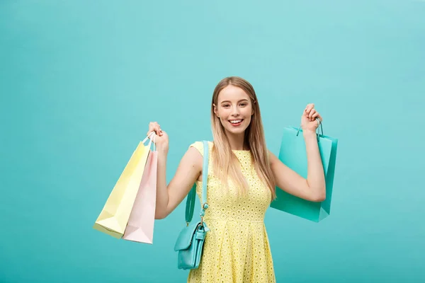 Életmód koncepció: portré megdöbbent fiatal vonzó nő sárga nyári öltözködés bevásárló táskák és nézi a kamera felett kék háttér. — Stock Fotó