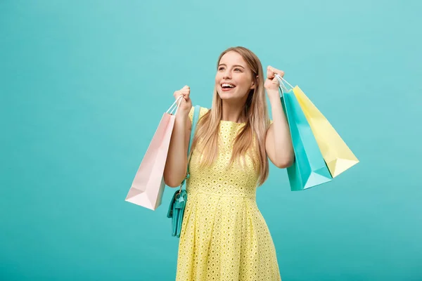 Lifestyle-Konzept: Porträt einer schockierten jungen attraktiven Frau in gelben Sommerkleidern, die mit Einkaufstaschen posiert und vor blauem Hintergrund in die Kamera blickt. — Stockfoto