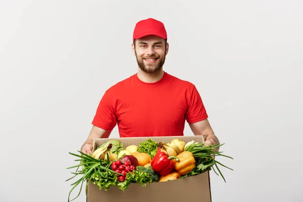 Teslimat kavramı - yakışıklı Cacasian teslimatçı bakkal yiyecek ve içecek deposundan paket kutu taşıyan. Arka plan gri studio izole. Kopya alanı.