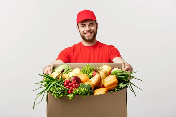 Teslimat kavramı - yakışıklı Cacasian teslimatçı bakkal yiyecek ve içecek deposundan paket kutu taşıyan. Arka plan gri studio izole. Kopya alanı.
