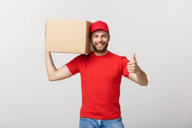 Neşeli, genç, yakışıklı teslimatçı elinde bir karton kutu ve gri arka planda dikilirken parmağını kaldırıyor.