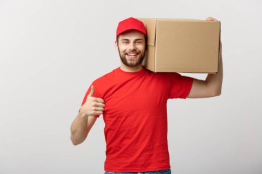 Neşeli, genç, yakışıklı teslimatçı elinde bir karton kutu ve gri arka planda dikilirken parmağını kaldırıyor.