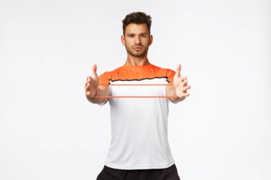 Motive edilmiş yakışıklı erkek sporcu spor aletleri kullanıyor, elleri ve kamerasıyla direnç bandı takıyor, fitness antrenmanları sırasında çömeliyor, beyaz arka plan