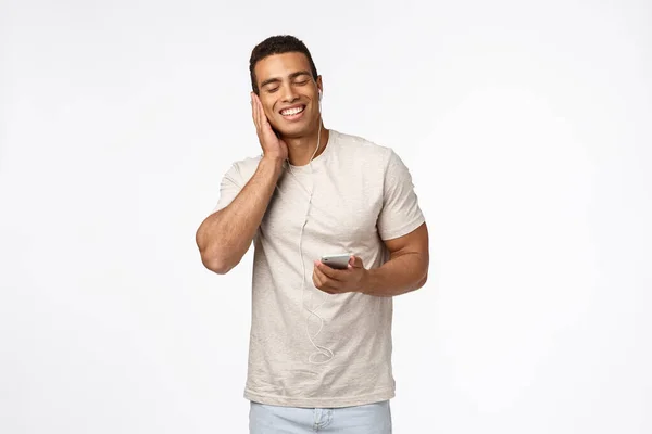 Feliz, hombre guapo complacido disfrutar escuchando música favorita, sosteniendo el teléfono inteligente, presione auriculares para oír y cerrar los ojos, sonriendo encantado, satisfecho con la calidad de los auriculares fantásticos, fondo blanco — Foto de Stock