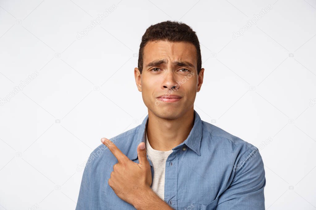 Skeptical, disappointed and doubtful handsome young man, frowning and grimacing from dislike, indecisive pointing left, cringe from reluctance, judging something unpleasant and bad, white background