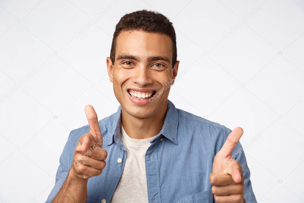 Cheeky, handsome charismatic hispanic man in blue shirt over t-shirt, pointing finger guns camera, smiling amused, encourage someone, meeting person with friendly greeting motion, white background