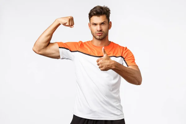 Sassy good-looking masculine bearded man in sports t-shirt, tense biceps, show muscles and thumb-up, make strong serious face, delighted with good body shape, productive workout, white background