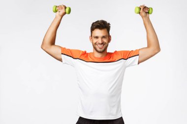 Spor ve spor salonu üyeliği tanıtımı. Spor kıyafetleri giymiş, kaslı, tatmin olmuş yakışıklı adam ellerini kaldırıyor, halterlerini sallayarak gülümsüyor, vücut şekliyle gurur duyuyor, plaj sezonuna uygun hale geliyor.