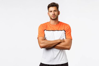 Spor eğitmeni, antrenmanı kaçıran müşteriyi hayal kırıklığına uğrattı. Yakışıklı, güçlü ve erkeksi bir adam, aktif giyimli bir futbolcu, spor tişörtü, göğsünde çapraz kollar, iddialı ve kararlı görünüyor.