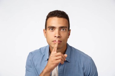 Ciddi görünümlü İspanyol genç adam kameraya doğru eğiliyor, şşşş yapıyor, şşşş yapıyor ya da şşşş yapıyor parmaklarını dudaklara bastırıyor, iddialı görünüyor, sessiz olun, sessizlik talep ediyor, beyaz arka plan
