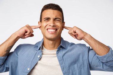 Üzüntülü mavi tişörtlü adam, rahatsız ve hoşnutsuzluktan suratını buruşturan, parmaklarıyla kulaklarını kapatan, sinen ve sıkılmış dişleri rahatsız eden, rahatsız edici yüksek ses duyan, beyaz arka plan