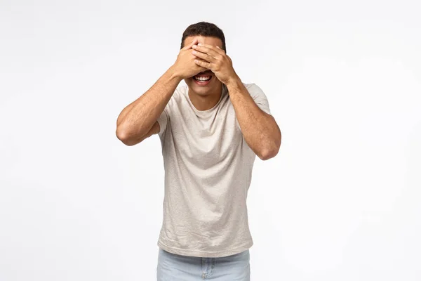 Handsome masculine young man playing hide-n-seek or peekaboo, cover eyes, standing blindfolded and smiling happy, waiting for signal to look at surprise christmas gift, white background