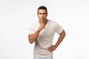 Ciddi görünümlü, güçlü, genç, kaslı bir adam sessizlik talep ediyor, sessiz ol, işaret parmağı dudakların yanında, kaşlarını çatma, sessiz ol, sessiz ol, kameraya sus, beyaz arka plan