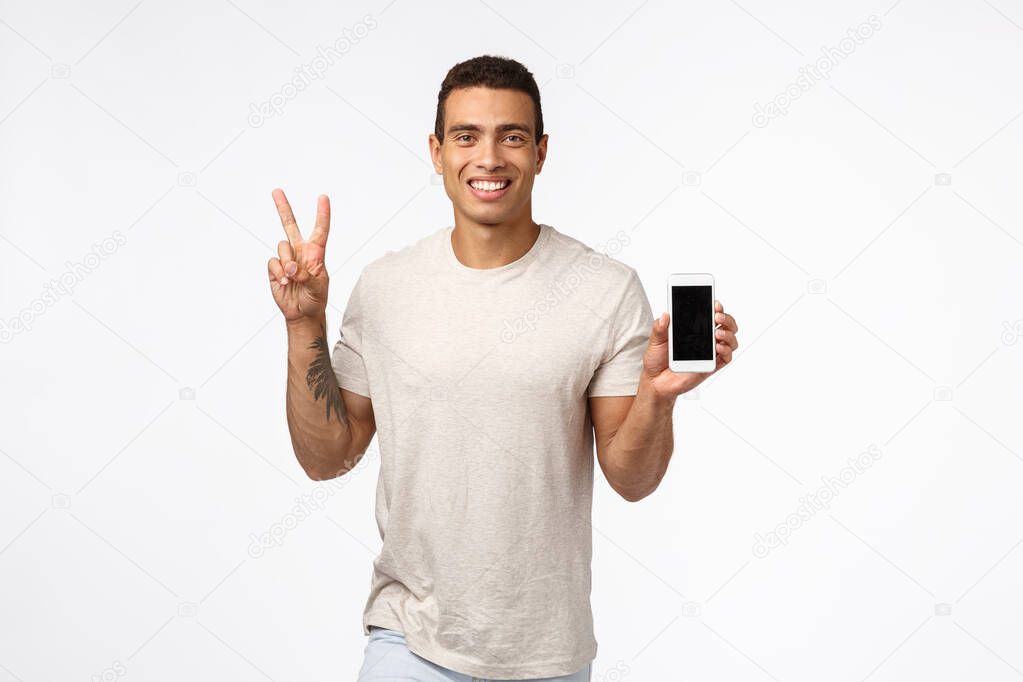 Friendly, cheerful handsome man in casual outfit, holding smartphone with blank screen, showing peace or victory sign, smiling, advertise site or app, recommend download mobile application