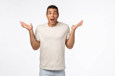 Beyaz tişörtlü yakışıklı, kaslı maço adam, şaşkınlık ve şaşkınlıktan ellerini yana çırpıyor, nefes nefese kalmış, kameraya nutku tutulmuş, beyaz arka plandan büyülenmiş.