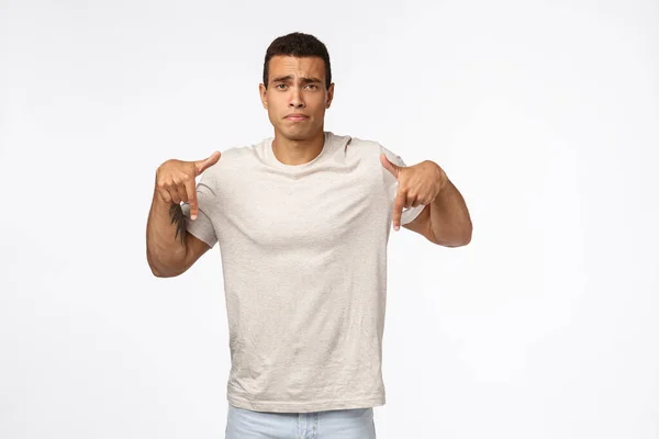 Decepcionado, sombrío joven bronceado hombre en camiseta, apuntando hacia abajo y mirando molesto, sintiéndose disgustado, angustiado por ver malos resultados, perder, exámenes fallidos, de pie fondo blanco deprimido — Foto de Stock