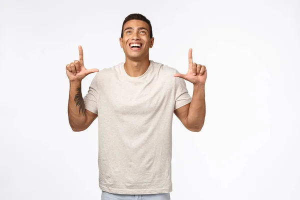Sexy hispânico muscular masculino no casual t-shirt, olhando para cima alegre, apontando topo propaganda, rindo e sorrindo de deleite e alegria, pé branco fundo divertido, ver algo bonito — Fotografia de Stock