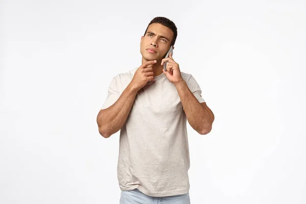 Thoughtful handsome young man in t-shirt, holding smartphone near ear, having conversation, touch chin and frowning, pondering choices, thinking how to answer, making decision, white background — Stock Photo, Image