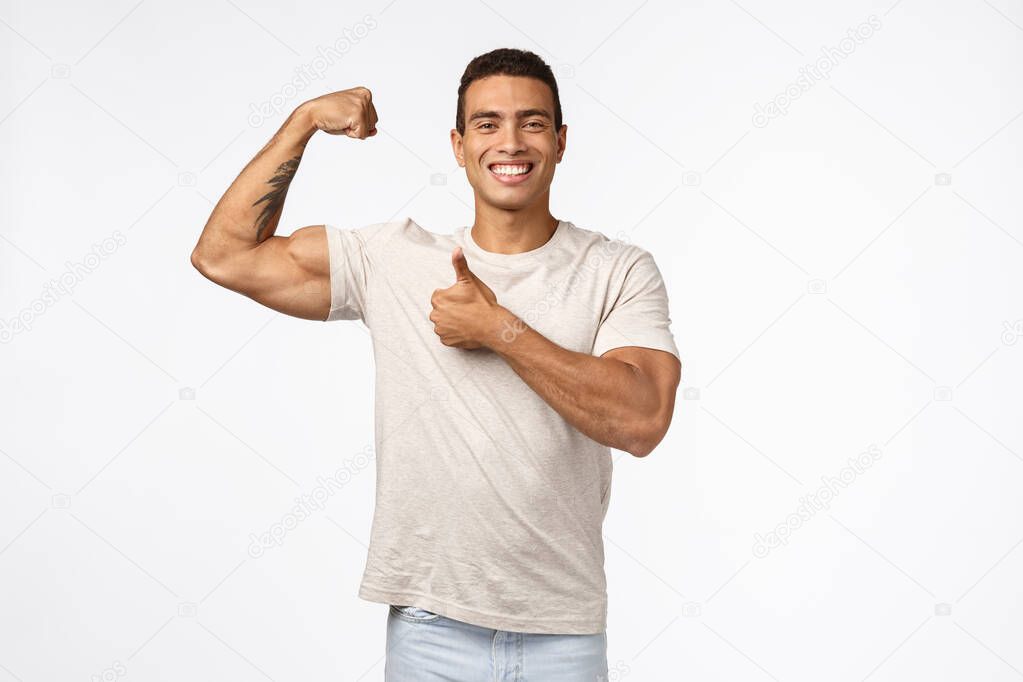 Handsome strong athletic sportsman in casual t-shirt, raise arm, tense muscle and proudly bragging good shape, big biceps, smiling pleased showing thumbs-up in approval and like, white background