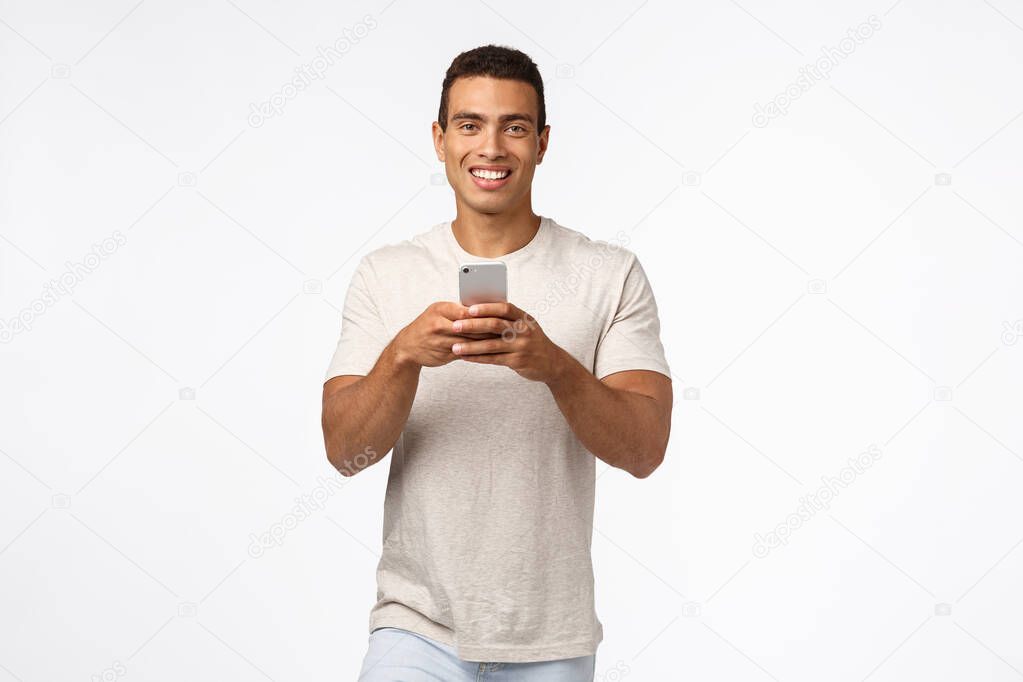 Technology, people concept. Handsome hispanic young man in t-shirt, holding smartphone, smiling joyful camera, browsing social media, download taxi app, shopping online, white background