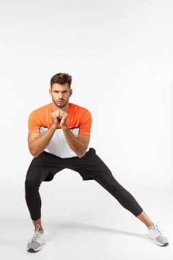 Dayanıklılık, motivasyon ve fitness konsepti. Ciddi görünümlü çekici sporcu yan çömelme hareketleri yapıyor, kollarını kenetliyor gerginlik, egzersiz, kas geliştirme, beyaz arka plan