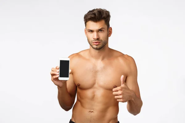 Sexy, cheeky handsome masculine young bearded man standing shirtless, holding smartphone, promote mobile app, make thumb-up in approval, recommend gym, show personal fitness rate on screen — Stock Photo, Image