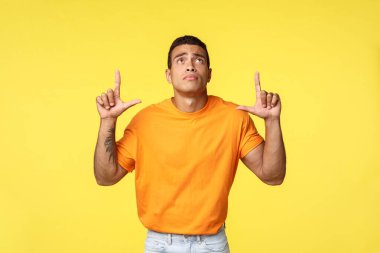 Turuncu tişörtlü, beyaz pantolonlu, üzgün ve üzgün bir genç adam. Üzüntü ve pişmanlıkla yukarıya bakıyor, iyi satışı kaçırıyor, bir şeyi kıskanıyor, sarı arka planda duruyor.
