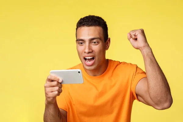 Радісний збуджений молодий красивий чоловік в помаранчевій футболці, піднімає руку на кулачковий насос як вкорінення для улюбленої команди, тримає смартфон горизонтально, дивиться футбол на мобільному пристрої, жовтий фон — стокове фото