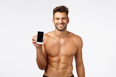 Teknoloji, reklam ve fitness konsepti. Seksi, güçlü, erkeksi, üstsüz atlet, elinde akıllı telefon, egzersiz uygulaması, spor salonu sosyal medya sayfası, sırıtan zevk, beyaz arka plan.