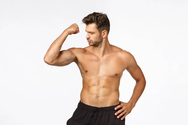 Charmant sportif caucasien avec corps masculin, debout torse nu, montrant des forces, a obtenu une forme parfaite, regardant serrer le muscle, montrant fièrement les biceps, debout fond blanc — Photo