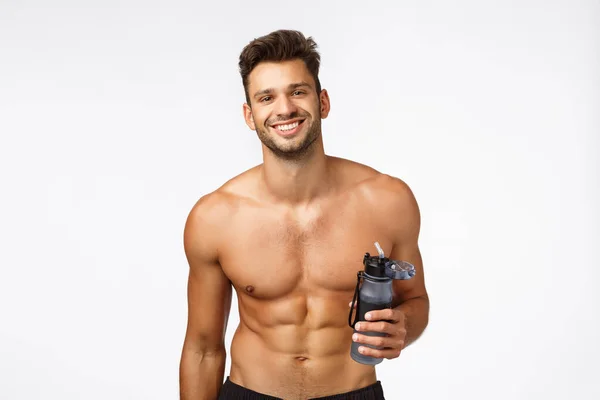 Fitness, health and workout concept. Handsome shirtless man with six-packs, perfect body, holding bottle, drink water after productive gym exercise, smiling happy and satisfied, white background — Stock Photo, Image