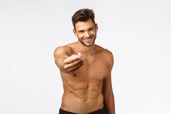 Sexy attractive smiling young shirtless sportsman give you one wireless earphone listen music together during workout, fitness training, grinning joyfully, standing naked torso show perfect body — Stock Photo, Image