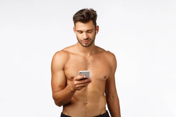 Technology, workout and fitness concept. Handsome shirtless strong sportsman put wireless headphones, hold smartphone smile as use workout app track down gym results, listen music during exercises — ストック写真