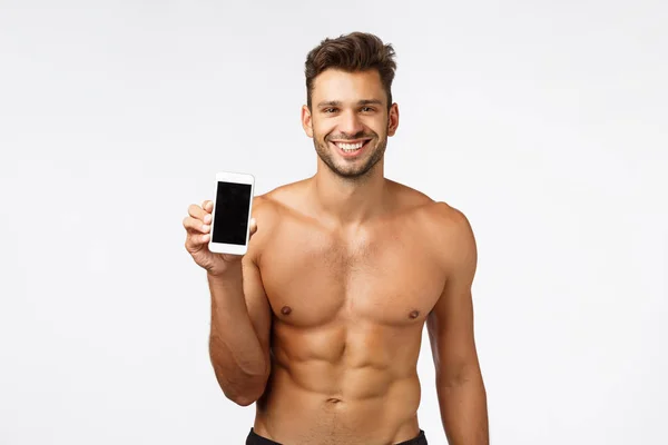 Technologie, inzerce a fitness koncept. Sexy úsměv silný, maskulinní atlet bez trička, držení smartphone, propagovat cvičení app, posilovna stránky sociálních médií, šklebící se potěšené, bílé pozadí — Stock fotografie