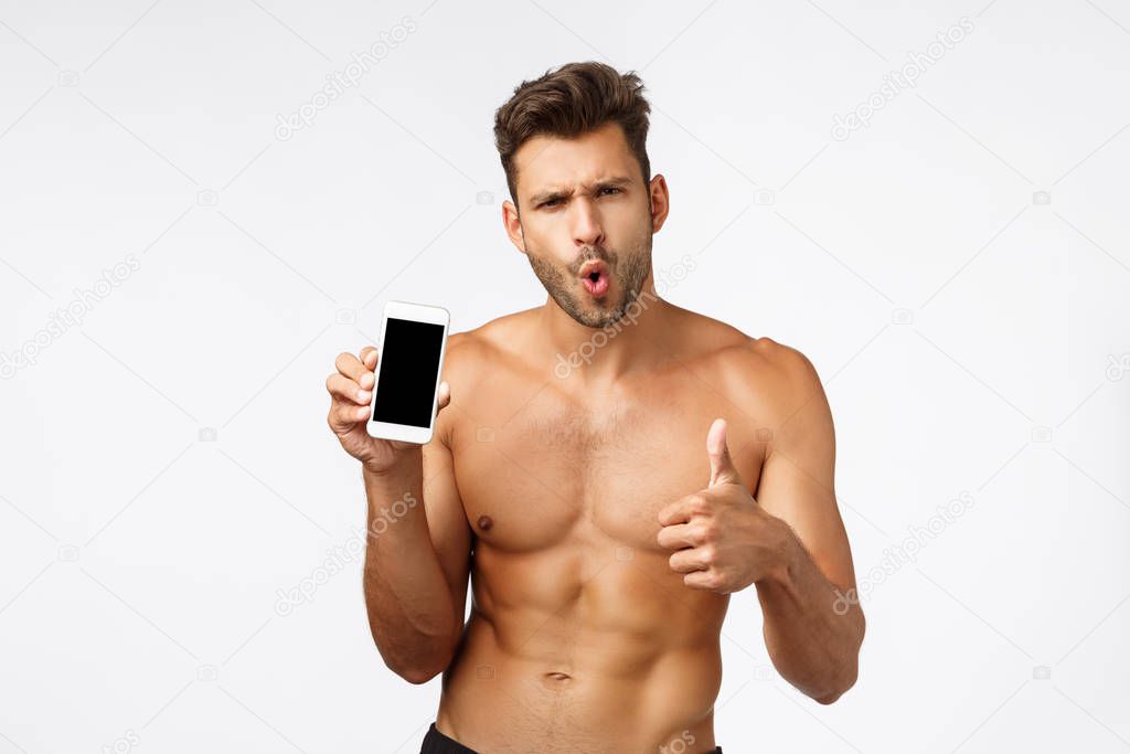 Astonished, impressed sexy young male athlete, sportsman with naked torso recommend good app, showing smartphone screen, folding lips from amazement and thumbs-up in approval, like application