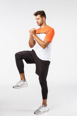 Şort, tayt ve spor tişörtü giyen yakışıklı, sakallı genç erkek atlet, kollarını sık, geril, spor salonunda egzersiz yap, ayağını kaldır, fitness egzersizleri yap, uzun beyaz arka plan