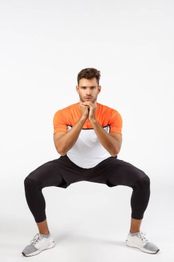 Aktif giyimli, uzun boylu, yakışıklı bir sporcu, ayakları yana aç, fitness egzersizi yap, kollarını sıkıca kavra ve otur, spor salonunun beyaz arka planı için poz ver.