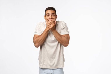 Yakışıklı, İspanyol tişörtlü, gülen ve gülümseyen genç kaslı adam sessiz kalmaya çalışıyor, kıkırdıyor, komik sahneler seyrediyor, beyaz arka plan.