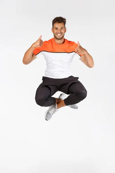 Teljes hosszúságú lövés férfi jumping, mozgás lövés boldog mosolygós sportoló a levegőben mutatja hüvelykujj-up jóváhagyása vagy hasonló, ajánlani félelmetes hely edzés, tornaterem promo, elégedett jó fitness berendezések — Stock Fotó