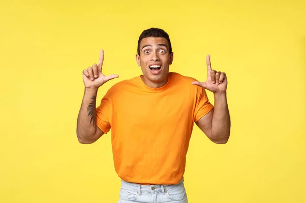 Fascinado, animado jovem caucasiano em t-shirt laranja, olhar câmera surpreendido e impressionado, apontando para cima como ver incrível oferta promo, de pé fundo amarelo surpreso . — Fotografia de Stock
