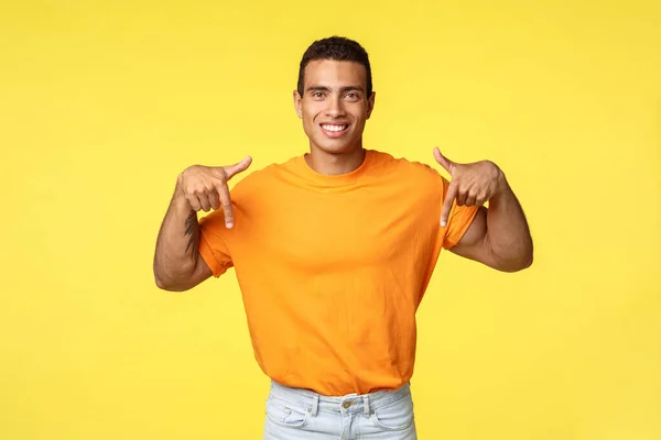 Hombre masculino joven guapo en camiseta naranja dando consejos donde comprar en línea, señalando sonriente asertivo y feliz, promover la publicidad, dar recomendación, fondo amarillo — Foto de Stock