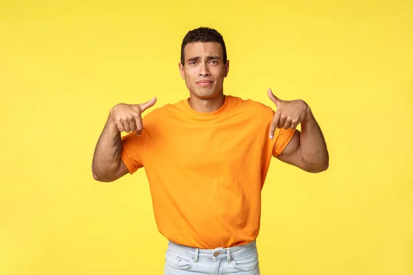 Bouleversé, triste mignon beau mec caucasien en t-shirt orange, pointant du doigt vers le bas, geste pour attirer l'attention mauvaises nouvelles, froncement de sourcils malheureux, sentiment de regret manqué chance, debout fond jaune — Photo