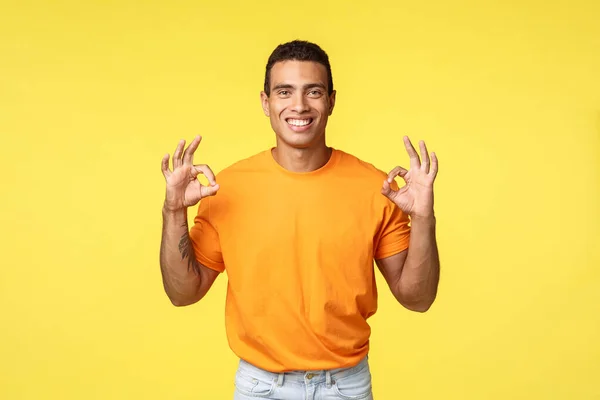 Encantador chico feliz con el brazo tatuado en camiseta naranja, pantalones blancos, mostrar bien, confirmación o buen gesto, sonriendo asentir de acuerdo, recomendar el producto, promoviendo la empresa en el fondo amarillo — Foto de Stock