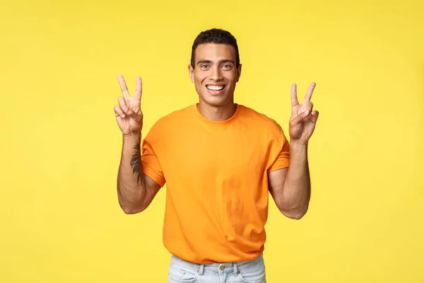 Guapo joven alegre en camiseta naranja, sonriendo optimista, hacer gesto de paz o citas signo, de pie fondo amarillo, expresar positividad y optimismo, de pie fondo amarillo — Foto de Stock