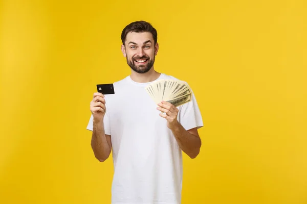 Young handsome happy smiling man holding banking card and cash in his hands isolated on yellow background. — Stock Photo, Image